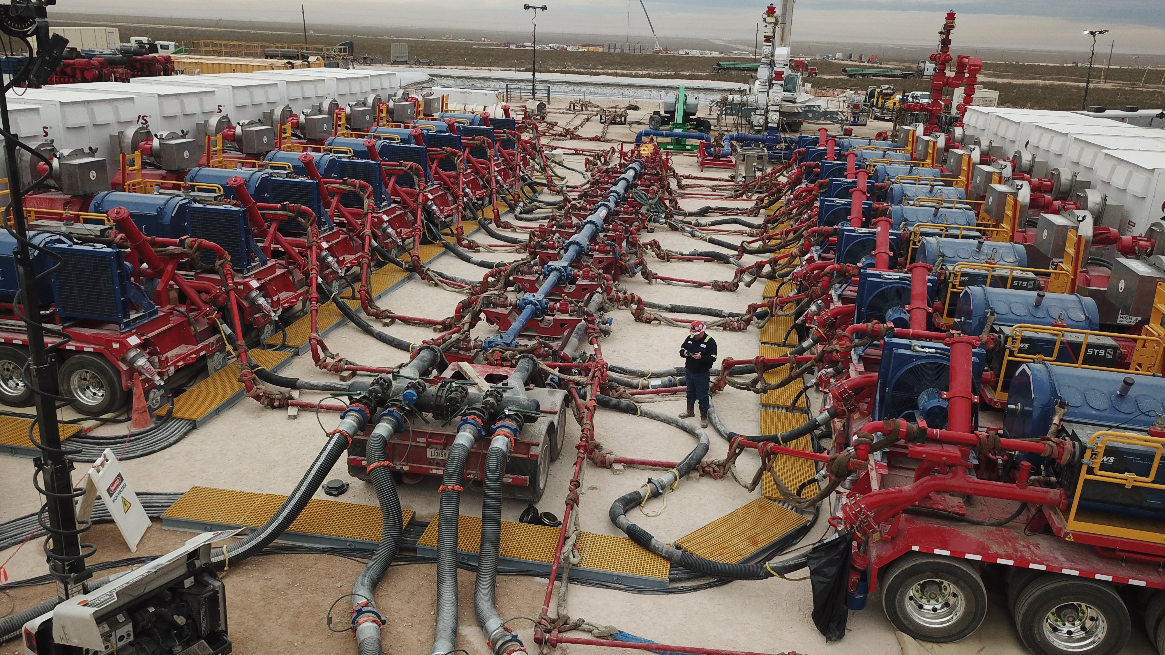 A large hydraulic fracturing site with numerous red and blue pumps, hoses, and other equipment arranged in a complex network, with a worker in protective gear standing in the middle.