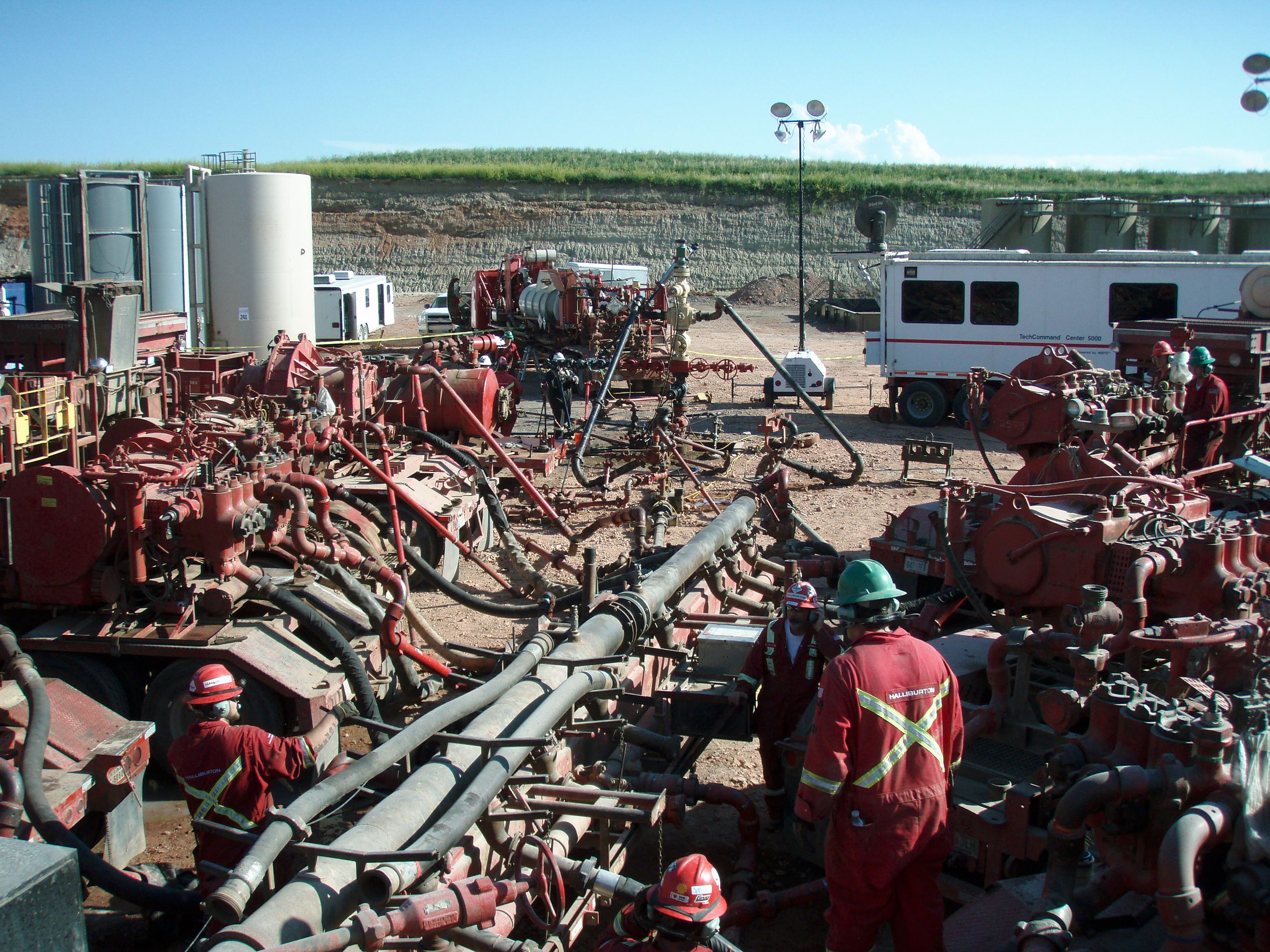 Workers in protective gear operate machinery at an industrial site with multiple pipes and heavy equipment.