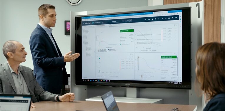 A man in a suit is giving a presentation with a large digital screen displaying charts and data, while two other people sit at a table with laptops.