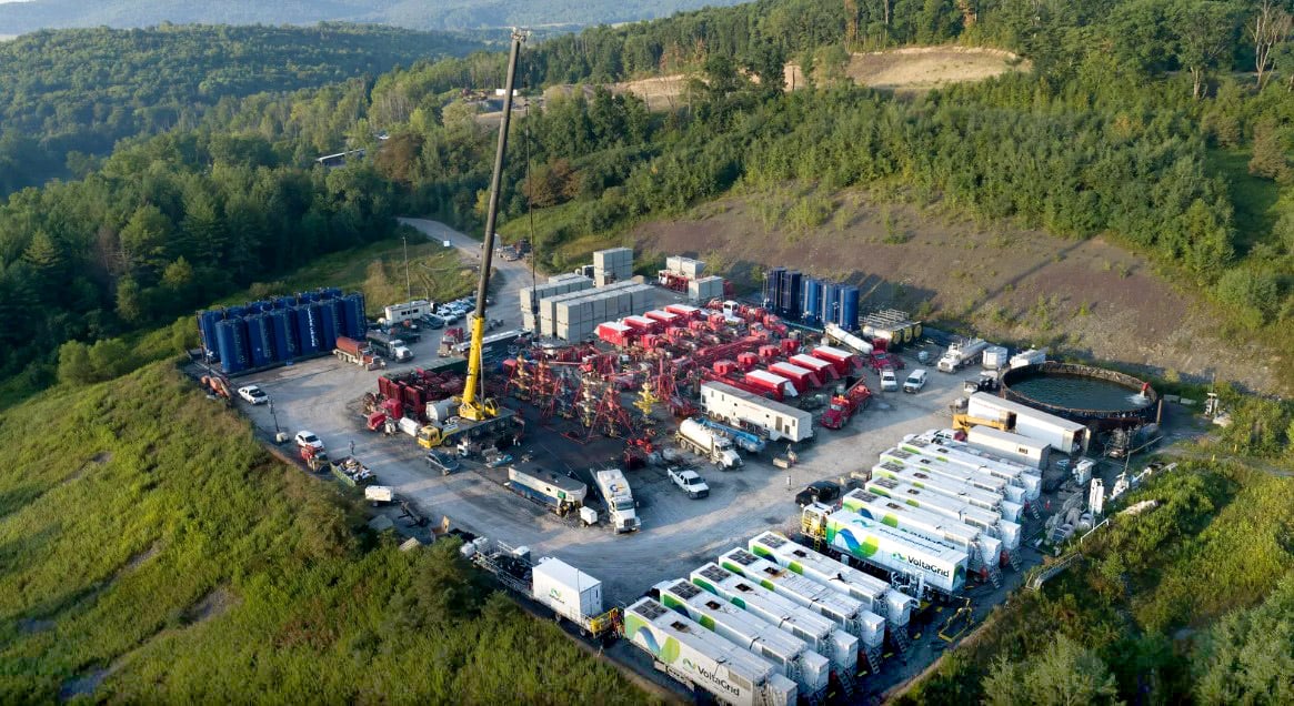 Aerial view of a fracking site surrounded by dense forest, featuring numerous trucks, large containers, and equipment arranged systematically on a cleared area.