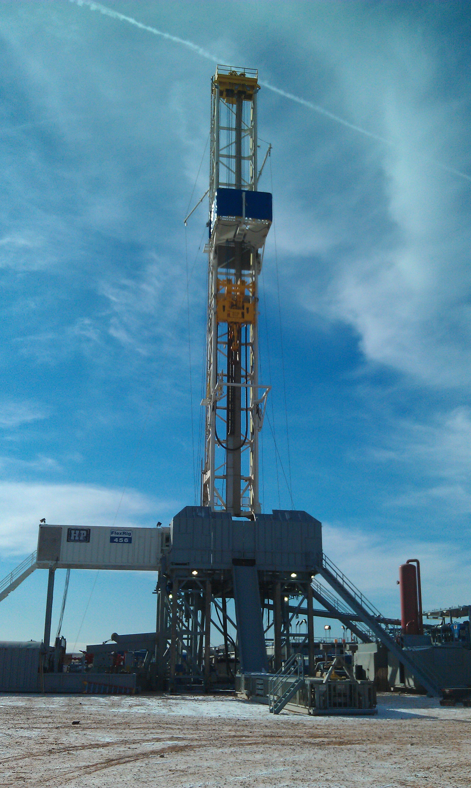 A tall oil drilling rig labeled FlexRig 456 stands against a partly cloudy blue sky.