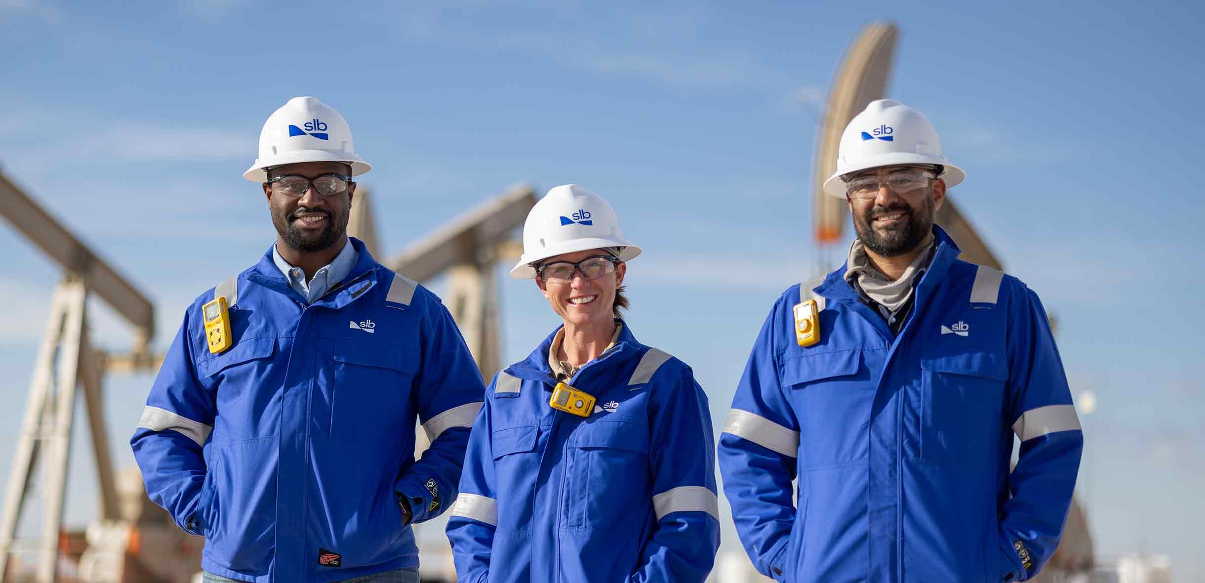 Three workers in blue jackets and white helmets stand outdoors, smiling, with an industrial facility in the background.