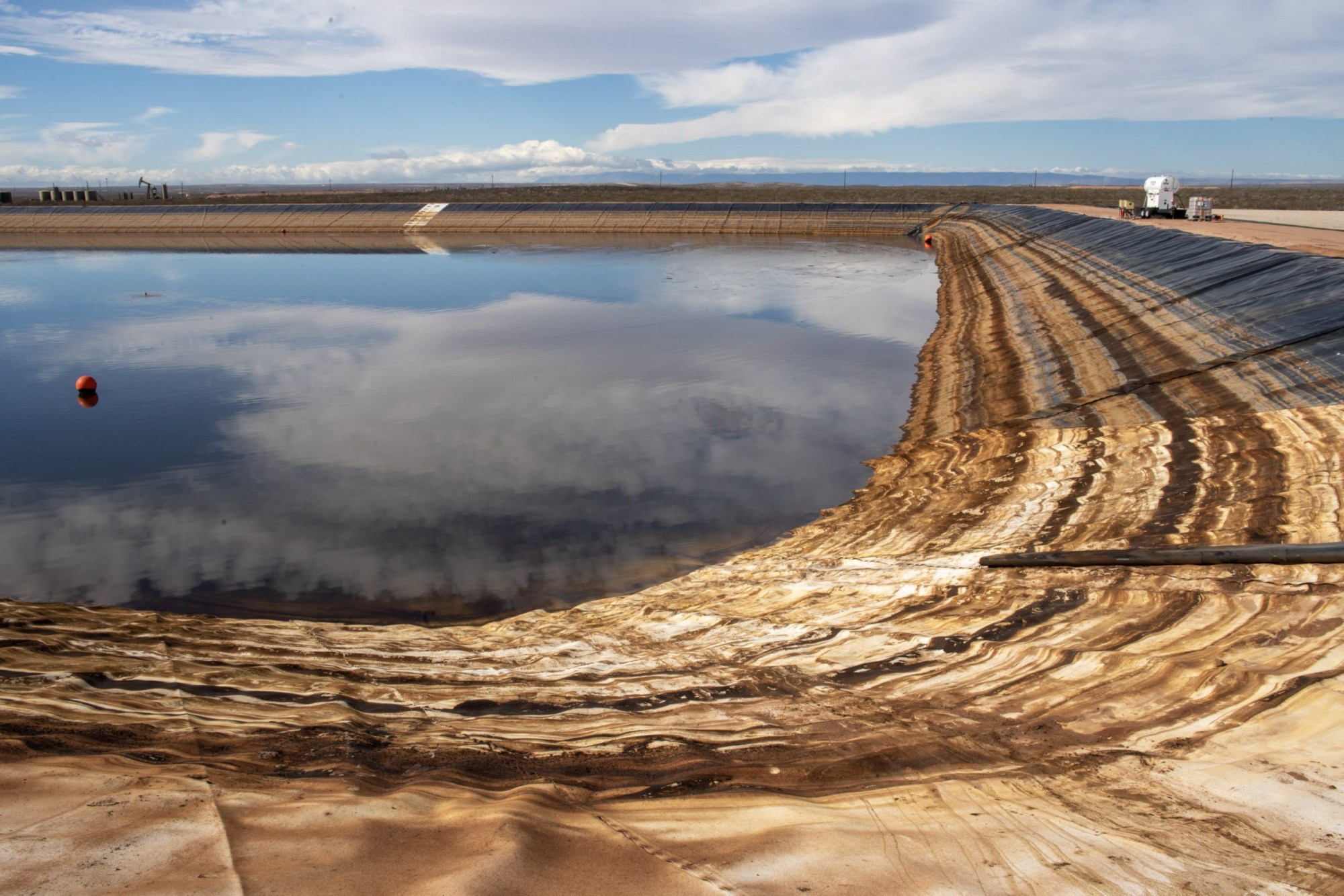 A large man-made water reservoir with a weathered plastic liner, reflecting a cloudy sky, with industrial equipment visible in the background.
