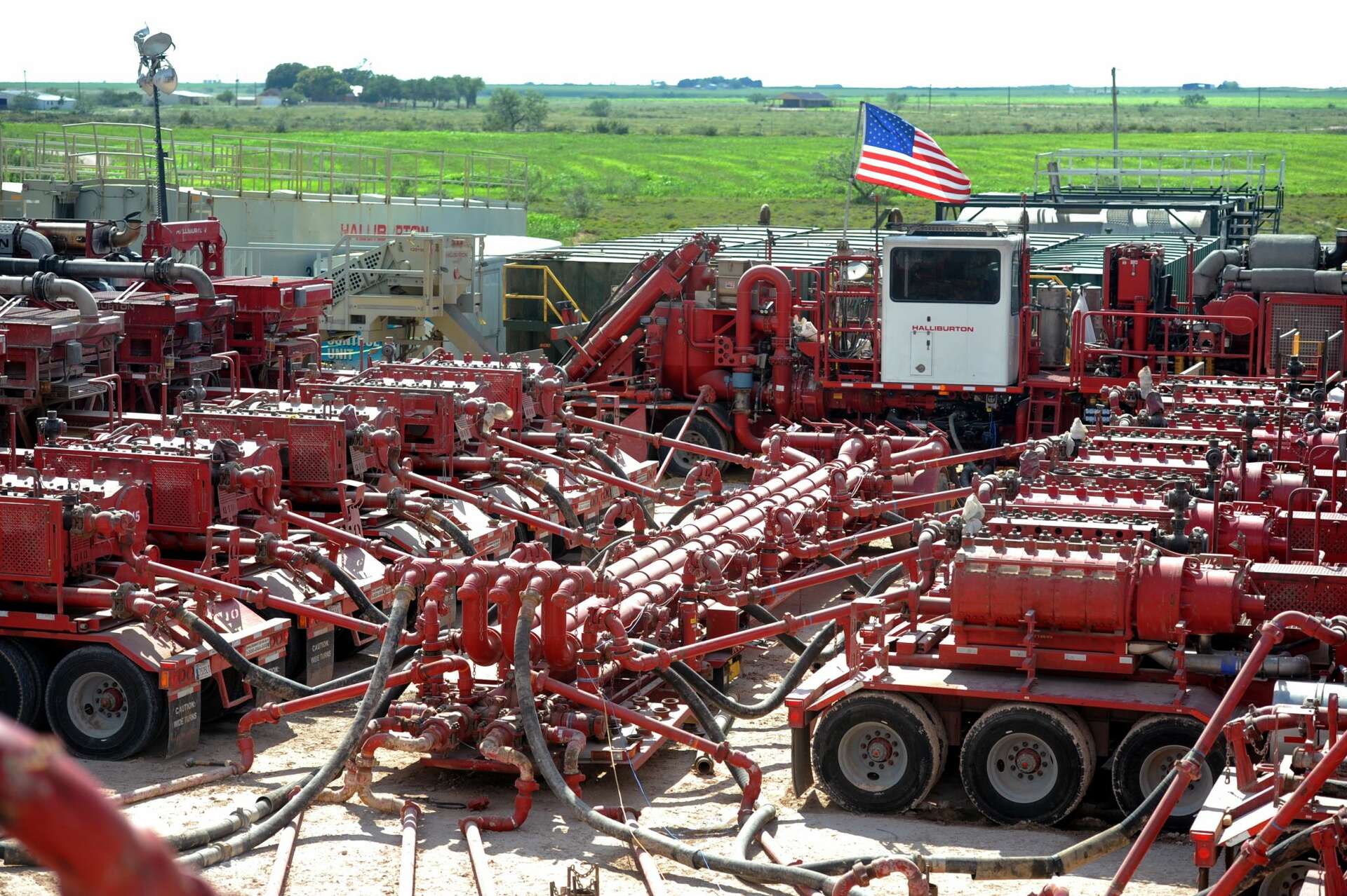 An oil field with a complex setup of red machinery and equipment connected by pipes, featuring an American flag in the background.