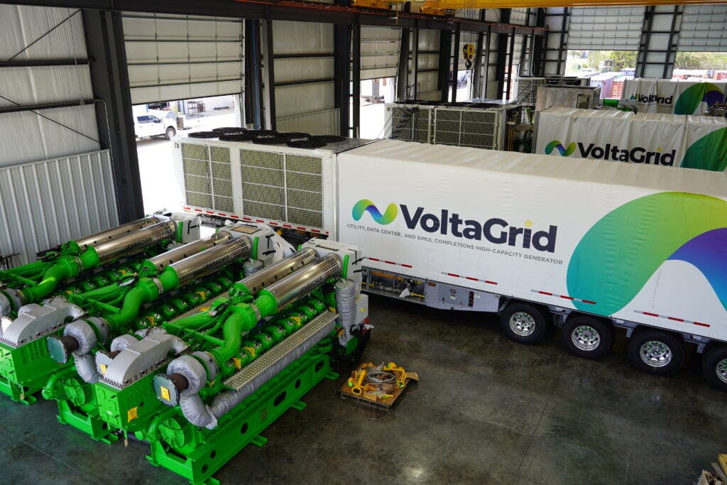 A warehouse with heavy industrial equipment, featuring a truck trailer labeled VoltaGrid and large green machinery.